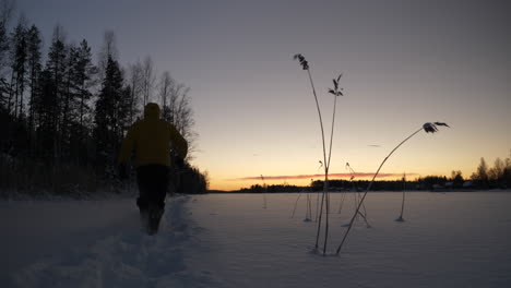 Man-walks-through-snow-towardss-winter-sunset,-static-shot