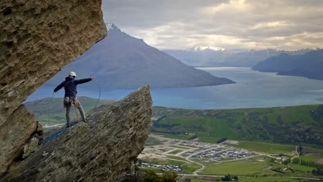 Mountain-rock-climber-coiling-rope-on-a-cliff-face-overlooking-a-beautiful-lake-during-sunset-on-a-cloudy-day
