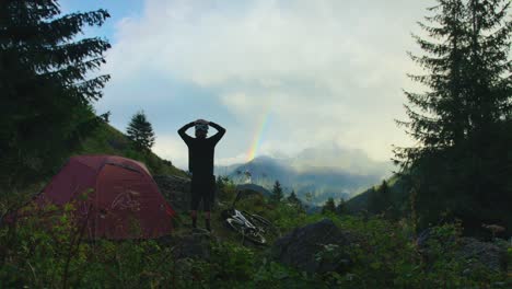A-cyclist-standing-by-his-campsite-is-looking-at-a-rainbow-in-the-distance