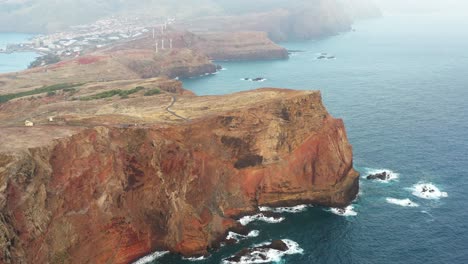 Aerial-View,f-Madeira-Island-Scenic-Coastline,-Steep-Cliffs-Above-Atlantic-Ocean,-Wind-Turbines-and-Canical-Town-in-Background,-Drone-Shot