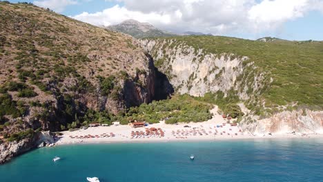 Vista-Aérea-De-Drones-De-La-Playa-Y-El-Cañón-De-Gjipe,-Dhermi,-Albania---Paraíso-Oculto-Con-Barcos,-Turistas,-Tumbonas-Y-Mar-Azul