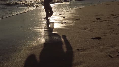 Shadow-of-carefree-young-boy-slow-motion-running-across-golden-wet-sand-beach-at-sunrise