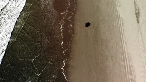 Aerial-view-of-a-lonely-black-car-driving-on-sandy-beach-during-golden-hours,-off-road-vehicle-between-ocean-waves-and-sand-dunes