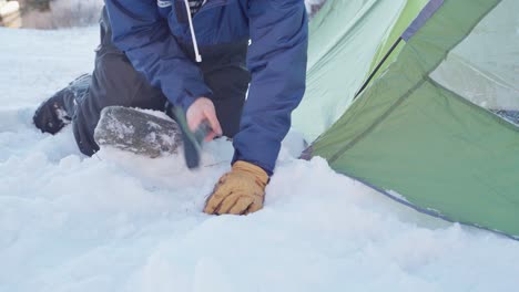 Camper-Hammering-The-Stake-On-Corner-Of-Camping-Tent-At-Winter-With-An-Axe-Then-Cover-With-A-Rock