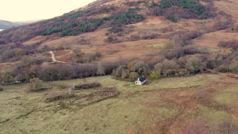 Slow-aerial-drone-footage-descending-towards-a-remote-Scottish-Bothy-,-fields,-long-shadows-and-a-forested-hillside-in-winter-with-a-backdrop-of-native-woodland