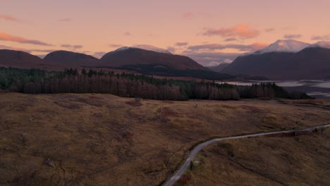 Aerial-drone-footage-flying-away-from-Glen-Etive-and-Loch-Etive-in-Scotland-during-an-orange-and-pink-sunrise-of-pastel-colours,-with-snow-capped-mountains-and-a-forest-in-the-background