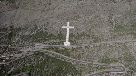 Drone-video-of-descriptive-panning-over-the-giant-white-cross-at-the-beginning-of-the-mountain-pass-of-the-Sh20-road-in-Bridje,-Albania,-houses-of-the-village-can-be-seen,-winding-road
