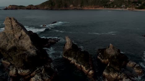 Aerial-rotating-shot-over-a-small-rocky-island-close-to-Mazunte-Beach,-Oaxaca,-Mexico-with-waves-crashing-on-the-island-at-sunset