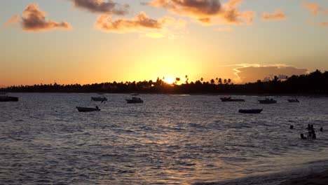 People-Swimming-In-The-Ocean-With-Boats-Floating-In-The-Distance-At-Sunset