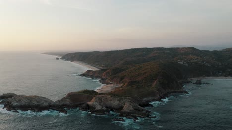 Epic-drone-shot-of-Mazunte-Beach-bordered-by-Punta-Cometa-,-which-is-a-small-peninsula-or-mountain-that-juts-out-from-the-shoreline,Mexico-during-sunset