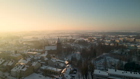 Luftbahnaufnahme-Des-Wunderschönen-Verschneiten-Stadtbildes-Mit-Kirche-Und-Verschneiten-Häusern-In-Polen-Während-Der-Sonnenuntergangszeit-Im-Winter