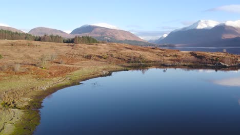 Aerial-drone-footage-flying-straight-up-to-reveal-Glen-Etive-and-the-shores-of-Loch-Etive-in-the-Highlands-of-Scotland-in-winter-with-snow-capped-mountains,-trees,-a-forest-and-still-reflective-water