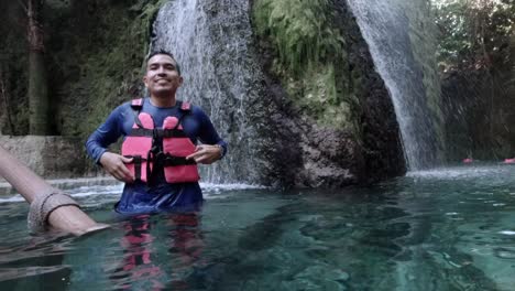Hispanic-Male-Speaking-wearing-a-Life-Jacket-in-a-Blue-River-with-Waterfall-in-the-Background