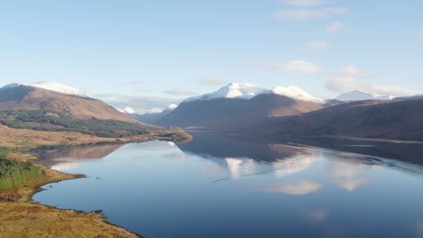 Backwards-flying-aerial-drone-video-revealing-an-epic-winter-view-of-Glen-Etive-and-Loch-Etive-in-the-Highlands-of-Scotland-with-snow-capped-mountains,-a-forest-and-beautiful-still-reflective-water
