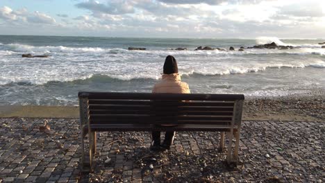 A-person-sits-alone-on-a-seaside-bench-watching-the-angry-waves-crash-ashore