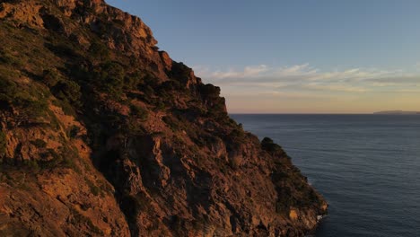 Drone-footage-of-a-sunset-lit,-jagged-looking,-seaside-cliff-along-the-coast-of-the-Mediterranean-Sea