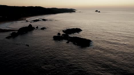 Aerial-tilt-up:-Silhouette-of-rocky-coastline-and-mountains-with-tranquil-Pacific-Ocean-during-sunset-in-backdrop