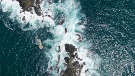 Tiny-fishing-boat-near-rocky-coast-while-strong-ocean-waves-hitting-it,-aerial-top-down-shot