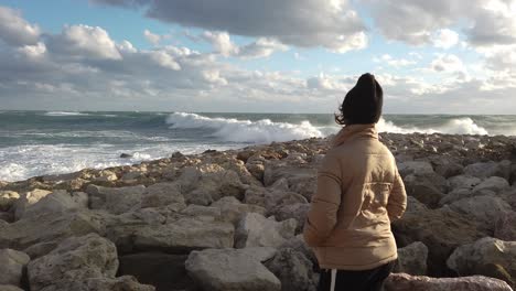 A-girl-stands-on-the-rocks-watching-the-large-waves-of-a-wild-sea-on-a-windy-day-in-Europe