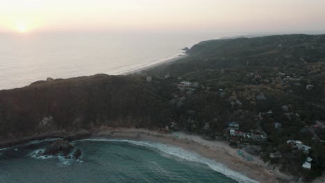 Toma-Aérea-De-Drones-De-La-Playa-Mazunte-En-La-Costa-Oaxaqueña-En-México-Con-La-Vista-De-La-Hermosa-Puesta-De-Sol-Sobre-El-Mar-En-El-Horizonte-Lejano