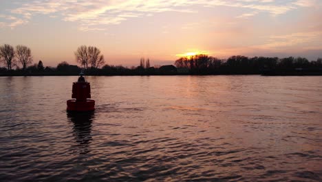 Silhouette-Der-Schwimmenden-Boje-Im-Fluss-Während-Gegen-Orange-Sonnenuntergang-Himmel-Mit-Lastkahn-Im-Hintergrund