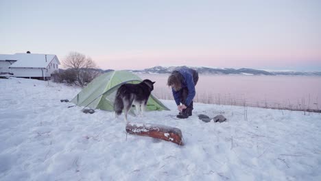 Camper-Mit-Alaskan-Malamute-Außerhalb-Des-Campingzeltes-Bei-Sonnenaufgang-Im-Winter