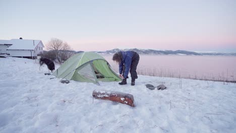 Mann-Bindet-Schnürsenkel-Aus-Schwarzen-Lederstiefeln-Auf-Verschneitem-Campingplatz-Am-Seeufer-Mit-Verspieltem-Alaskan-Malamute,-Der-Bei-Sonnenuntergang-Herumläuft