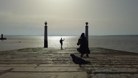 Person,-Die-Foto-Von-Posierendem-Freund-Macht,-Tejo-Im-Hintergrund,-Silhouettenrückansicht,-Cais-Das-Colunas,-Lissabon,-Portugal