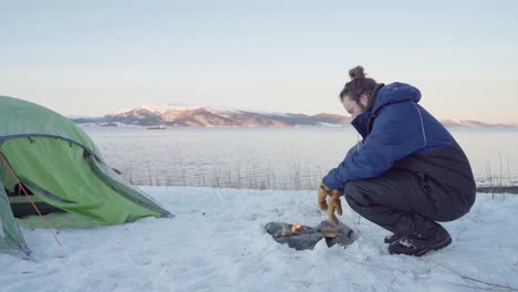 Man-In-Winter-Clothing-Sitting-By-The-Fire-Outside-Camping-Tent-In-Snow-At-Daytime