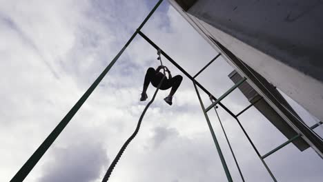 Dynamic-slow-motion-footage-of-a-fit-woman-descending-after-a-successful-rope-climb,-backlit-against-the-clouded-sky