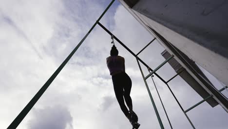 Dynamic-slow-motion-footage-of-a-fit-woman-succeeding-at-reaching-the-top-while-climbing-a-rope,-backlit-against-the-clouded-sky