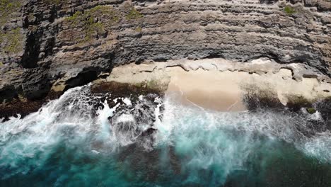 Aerial-view-of-waves-rolling-onto-the-beach-below-a-cliff-in-Uluwatu,-Bali
