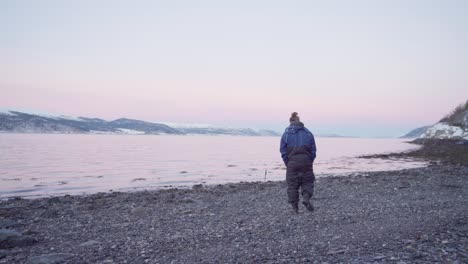 A-Man-In-Winter-Clothing-With-His-Dog-Walking-On-Pebbled-Shoreline-Near-Vanvikan,-Indre-Fosen,-Norway