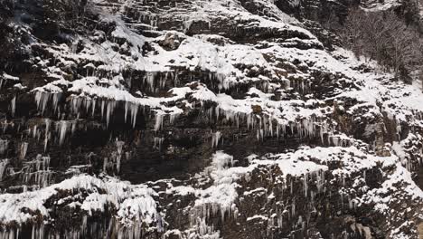 An-Aerial-View-of-Cirque-du-Fer-à-Cheval-while-covered-in-snow-during-a-cold-winter,-close-up-on-a-cliff-face-covered-in-ice-stalactites-with-a-slow-upwards-movement