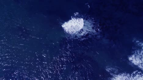 Blue-ocean-water-with-rocks-visible-below,-top-down-drone-shot