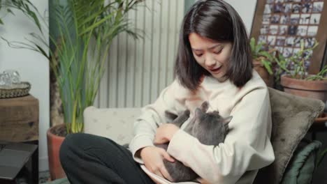 Black-haired-Asian-woman-sits-on-the-couch-and-strokes-her-cat-on-her-head-while-hugging-and-kissing-it
