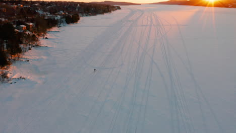 Scenic-sunset-aerial-of-a-frozen-lake-as-a-person-snowshoes-along-the-ice-and-snow