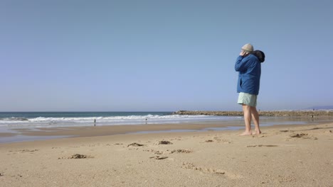 Older-Male-Taking-Phone-Call-On-Beach-Wearing-Blue-Coat
