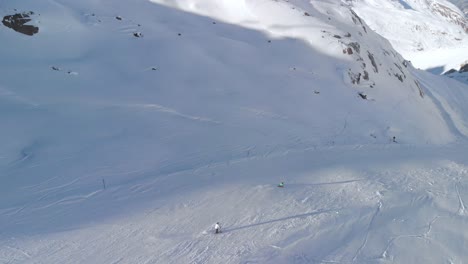 Aerial-view-following-slalom-skiers,-riding-downhill-on-the-slopes-of-the-Kaunertal-alps,-in-Austria---pan,-drone-shot