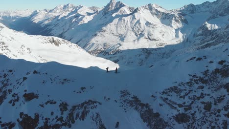 Aerial-view-over-mountaineers,-with-skis-on-top-of-a-mountain-ridge,-sunny,-winter-day-in-the-alps---pull-back,-drone-shot