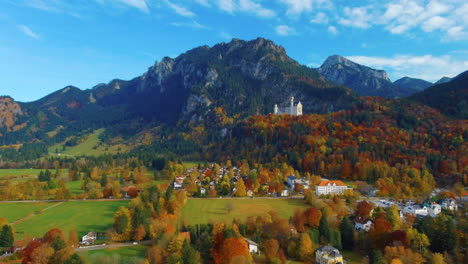 Relaxing-view-of-drone-lowering-down-towards-village-houses-towards-castle-on-hill-over-scenic-autumn-field-in-the-afternoon-near-the-Neuschwanstein-Castle-in-Germany,-Europe,-wide-view