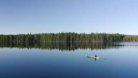 Hombre-En-Kayak-Verde-Rema-Tranquilamente-A-Través-De-Un-Lago-Muy-Suave-Que-Refleja-El-Borde-Del-Bosque-Entre-La-Hermosa-Naturaleza-De-Finlandia-En-Un-Día-Soleado