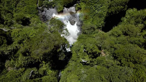 Aerial-lowering-on-Salto-El-Leon-waterfall-with-mist-and-rainbow-surrounded-by-green-dense-forest,-Pucon,-Chile