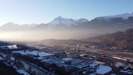 Drone-flight-against-the-light-fog-on-the-horizon-over-the-town-of-Sion-in-the-Swiss-Valais-in-sunny-weather-and-snow-capped-mountains