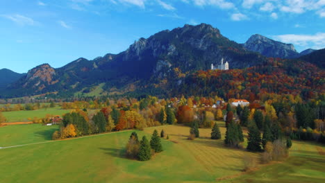Curved-angled-shot-of-drone-gliding-and-flying-over-village-houses-towards-castle-on-hill-over-scenic-autumn-field-in-the-afternoon-near-the-Neuschwanstein-Castle-in-Germany,-Europe,-wide-view