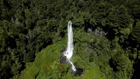 Aerial-dolly-in-lowering-on-Salto-El-Leon-waterfall-falling-into-rocky-pool-surrounded-by-green-woodland,-Pucon,-Chile