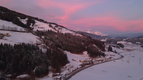 Aerial-shot-of-a-snow-covered-mountain-town,-winding-road-through-the-valley-during-sunset