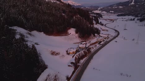 Aerial-shot-of-a-snow-covered-mountain-town,-winding-road-through-the-valley-during-sunset