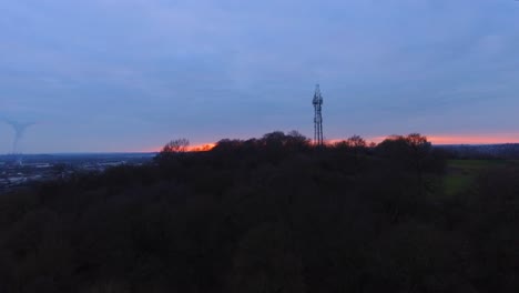 Telephone-mask-on-top-of-a-hill-surrounded-by-trees-and-woodland-at-sunset-dusk-moving-in-drone-aerial-shot