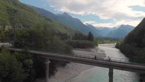 aerial-side-view-of-two-cyclists-crossing-bridge-across-clear-alpine-river,-summer